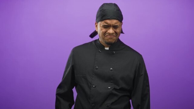 Hispanic man chef grimaces, face and beard scrunched, wearing black chef uniform and bandana in studio; disgust unease.