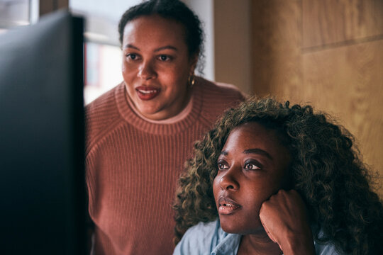 Focused curly haired businesswoman analyzing data on computer with female coworker in tech office