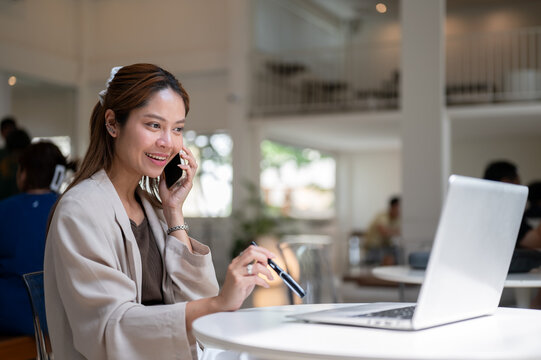 Pretty asian woman holding pen talking on phone call in front of laptop as sitting at table in cafe.