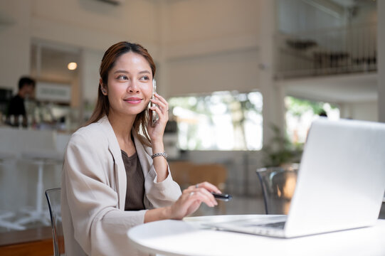 Smiling asian woman holding pen listening to phone call in front of laptop sitting at table in cafe.