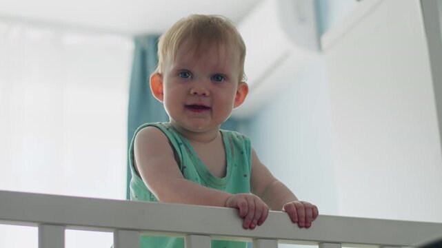 Baby girl holding crib railing and learning to keep balance. Baby milestones first steps