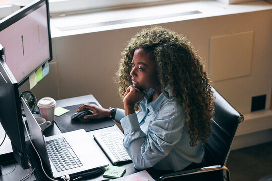 High angle view of concentrated female business professional analyzing data preview on dual monitor in startup office