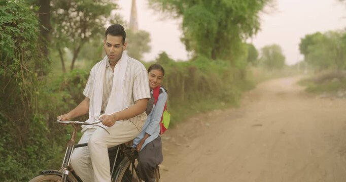 Indian happy young adult dad talk speak drop school to teen girl child rural area place road path way. Smiling female wear uniform with bag sitting on cycle back seat enjoy ride have fun joy desi life