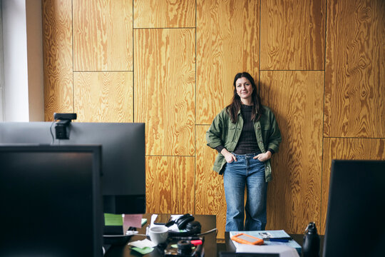 Portrait of confident female tech entrepreneur leaning on wall with hands in pockets at startup office