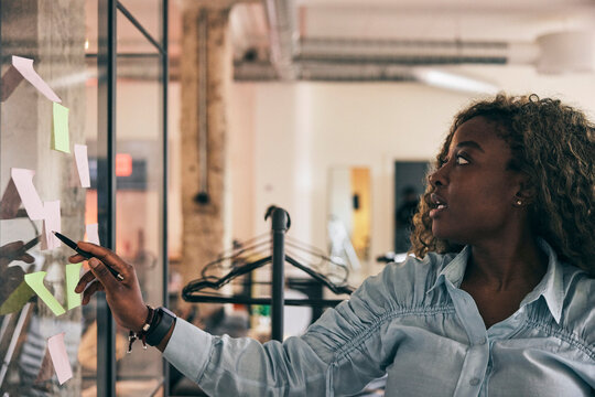Curly haired female business professional writing ideas on adhesive notes stuck on glass wall in startup office