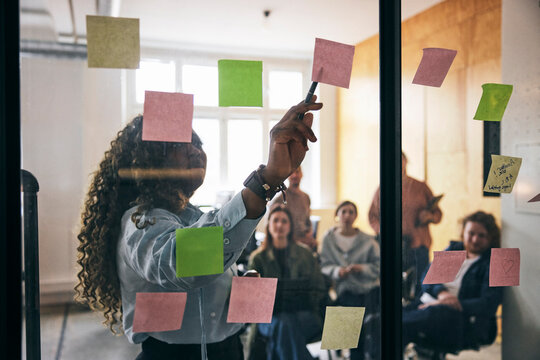 Curly haired female entrepreneur explaining points through adhesive notes to team in startup office meeting