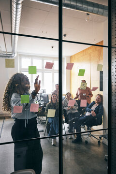 Curly haired female business expert explaining points through adhesive notes to team in startup office meeting