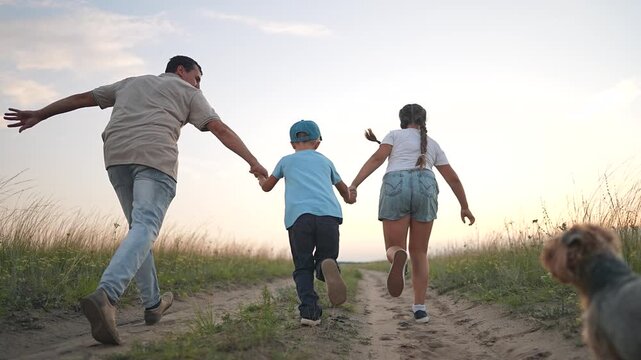 Father runs with children holding hands on dirt path at sunset with dog following. Man with boy and girl play outdoors. Children run with father and terrier. Family enjoys sunset in countryside field.
