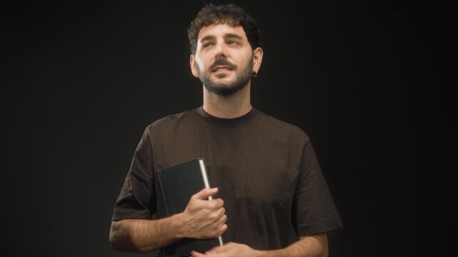 Man with beard and brown t shirt clutching black hardcover book with hand to chest in studio under soft lighting; quiet contemplation.