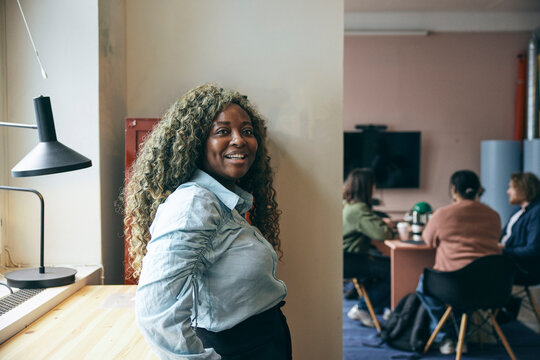 Portrait of happy female business entrepreneur with curly hair standing near cabinet in startup tech hub