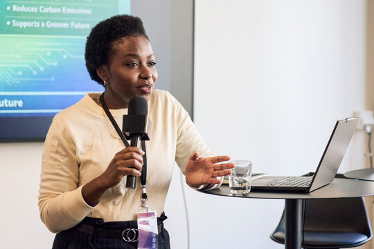 Professional female speaker holding microphone gesturing while giving presentation at seminar in convention hall