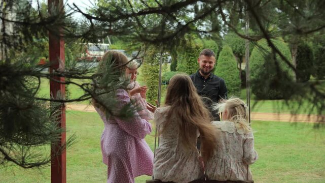 Young father pushing his two daughters on a garden swing while the mother holds another child nearby, creating a heartwarming scene of familial bonding and outdoor fun in a lush green park