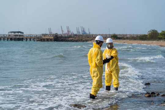 Environmental engineers in hazmat suits collecting and inspecting water samples near industrial gas storage tanks for pollution and safety monitoring.