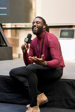 Happy male presenter communicating through microphone while sitting on platform during presentation speech in convention