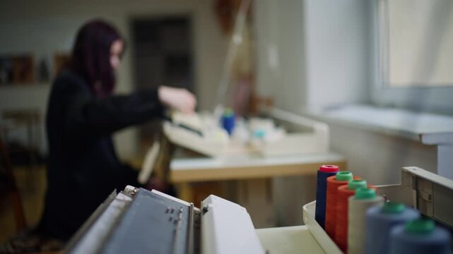 A blurred shot of female artisan works on knitting machine, focus on the foreground where colorful thread spools stand in a row near window.