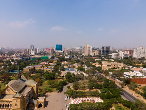 Aerial view of Frere Hall surrounded by lush green trees and the modern city skyline under a clear blue sky in Karachi, Sindh, Pakistan.