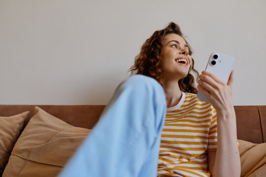 Joyful woman relaxing at home on a sofa, looking at a smartphone, striped top, soft cushions, natural light, casual lifestyle moment, cozy interior