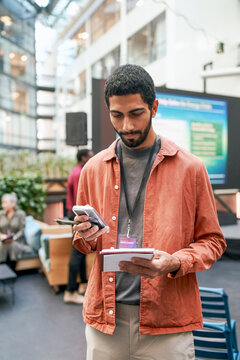 Male entrepreneur cross-referencing information with smart phone and notepad while standing in convention center