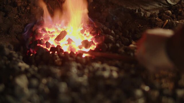 Close-up of a man&rsquo;s hand turning a metal rod in flames among charred rocks and coal, showcasing blacksmithing, craftsmanship, and dramatic forge action.