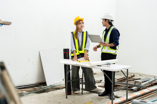 Asian woman engineer and male construction supervisor discuss report near blueprint table, planning unfinished building renovation, safety compliance, quality inspection, contractor coordination.
