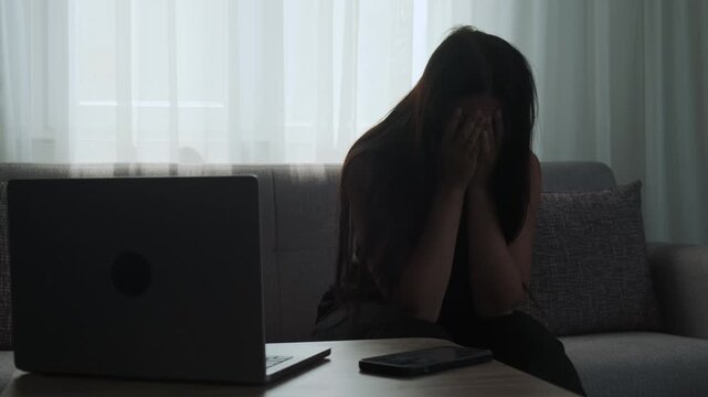 Depressed woman with head down sitting on sofa in despair at home next to laptop and phone, illustrating mental fatigue. Realistic depiction of freelance burnout