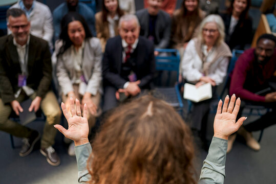 High angle view of female speaker gesturing with open palms toward diverse audience seated in convention hall