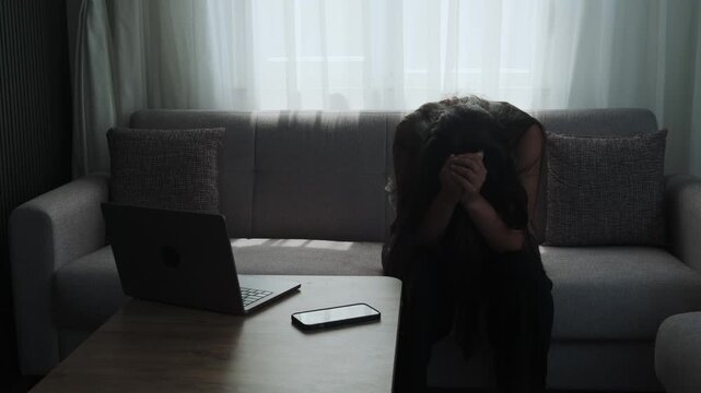 Depressed woman with head down sitting on sofa in despair at home next to laptop and phone, illustrating mental fatigue. Realistic depiction of freelance burnout