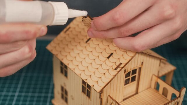 Constructing wooden house model with hands attaching chimney piece using glue during DIY miniature home assembly. Real estate concept, creative architecture hobby, handmade housing prototype on