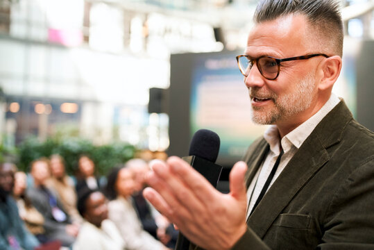 Mature male keynote speaker speaking into microphone at business seminar in convention hall