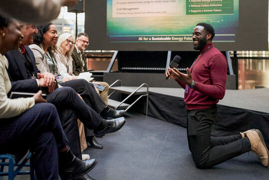 Full length of smiling male speaker kneeling while communicating with audience during business presentation at conventio