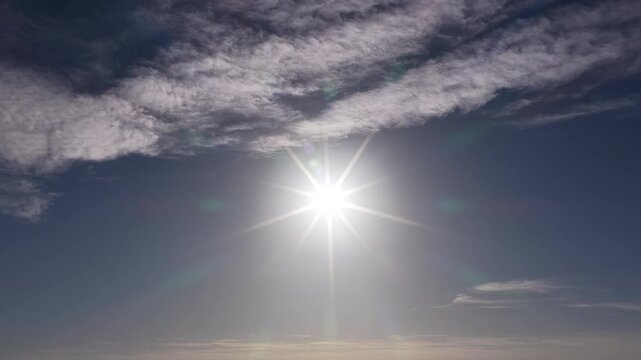 Bright sun with starburst rays shining through scattered clouds in blue sky