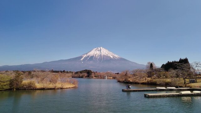 Picturesque wide angle view of Mount Fuji towering over a calm lake, featuring tourists appreciating the scenery from a dock.