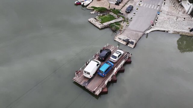 Stationary overhead drone shot of the wooden cable ferry carrying cars as it approaches the concrete docking ramp on the Vivari Channel shore in Butrint, Albania
