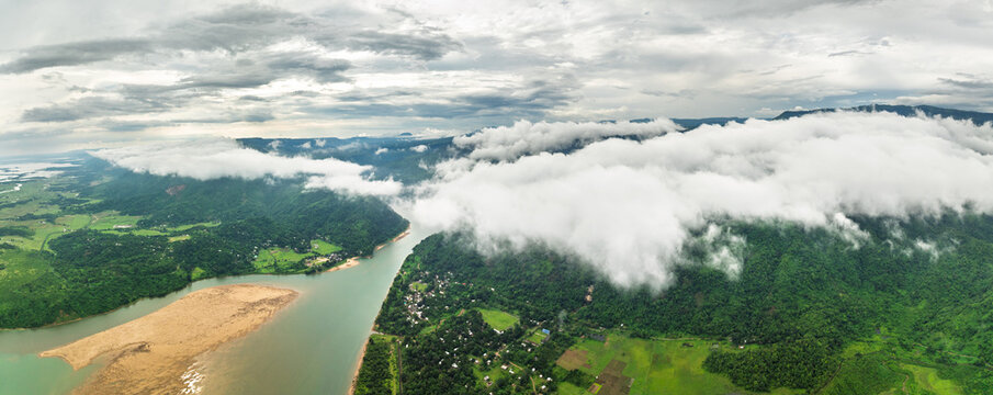 Aerial view of the winding Jadukata River flowing through lush green hills under a thick layer of white clouds in Sunamganj, Sylhet Division, Bangladesh.