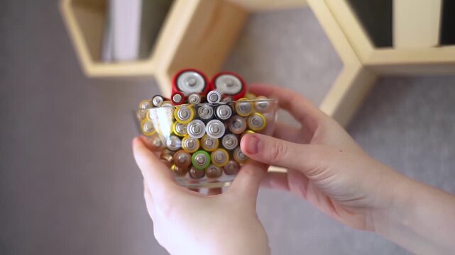 A young woman places a glass bowl filled with used batteries onto a wooden shelf for future disposal. This process demonstrates conscious household waste sorting and care for the environment.