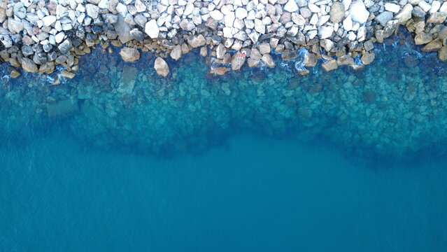 Top down aerial drone view of transparent turquoise sea water and rocky shoreline with a lone fisherman