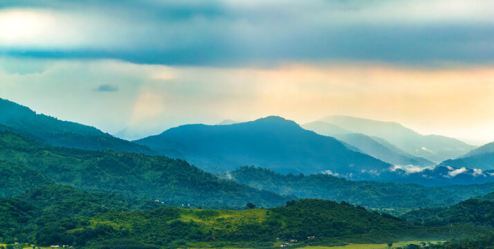Aerial view of lush green rolling hills and misty mountain peaks under a dramatic cloudy sky in the rural landscape of Sunamganj, Sylhet Division, Bangladesh.