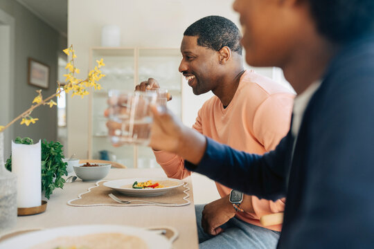 Happy man holding glass of water while sitting with son at dining table during lunch time