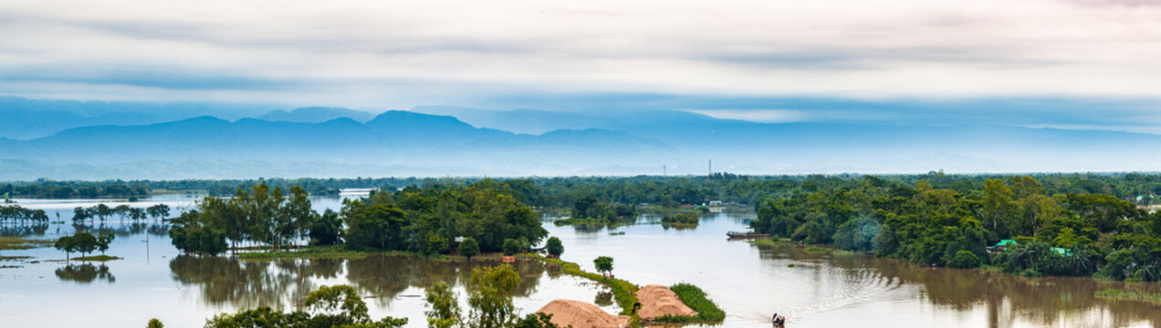 Aerial view of the flooded landscape with lush green trees, sand piles and distant blue mountains under an overcast sky in Sunamganj, Sylhet Division, Bangladesh.