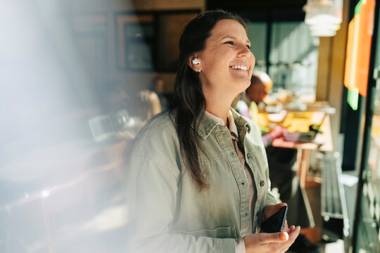Happy businesswoman looking away while talking on call using wireless earbuds at office
