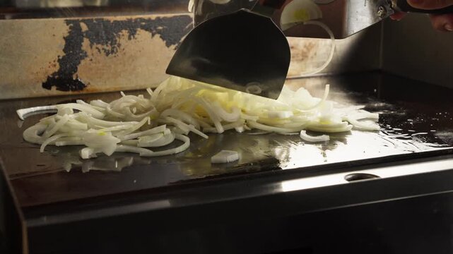 a person cooking thinly sliced yellow onion in hot flat griddle