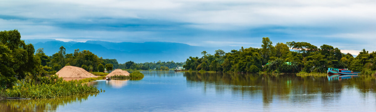Aerial view of a wide river flowing past lush green trees and sand piles with blue mountains under a cloudy sky Sunamganj, Sylhet Division, Bangladesh.