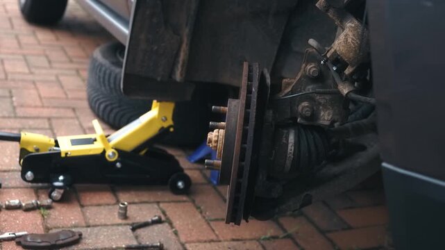 Raised car on jack with front wheel assembly turning, exposed brake disc rotor and suspension during vehicle repair. Automotive maintenance concept featuring wheel hub, brake system components