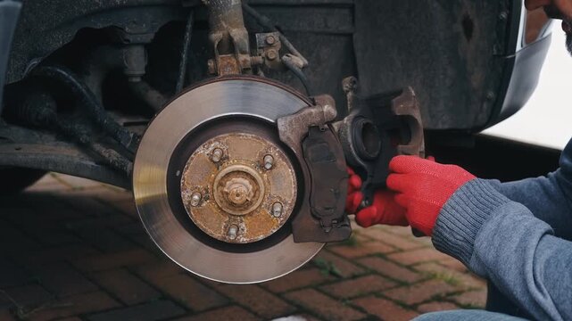 Removing brake caliper assembly from car disc brake system. Repairing vehicle braking mechanism with hands detaching caliper from rotor hub outdoors. Automotive maintenance process focusing on brake