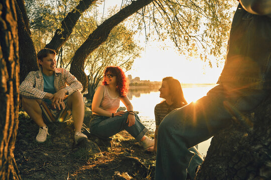 Friends resting by riverside trees at sunset with nostalgic film grain. Concept of slow living, summer memories, youth culture, weekend escape, travel blogs, and peaceful social bonding.