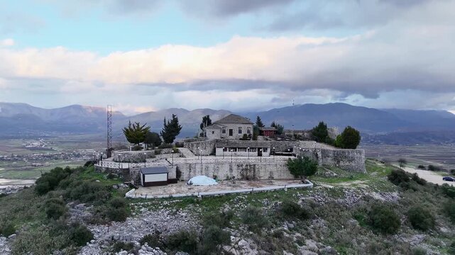 Aerial tracking shot pulling back from the stone terraces, central restaurant building, and fortification walls of Lekuresi Castle overlooking a vast mountain valley in Albania