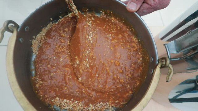 Macro shot of a person stirring a viscous, sweet molasses and nut mixture in a pot during traditional food preparation.