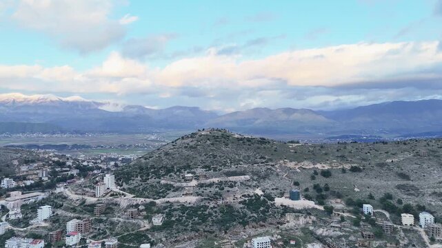Aerial tracking shot moving forward over a rugged green ridge toward the hilltop Lekuresi Castle complex, revealing distant snowy mountain ranges under a cloudy sky in Albania