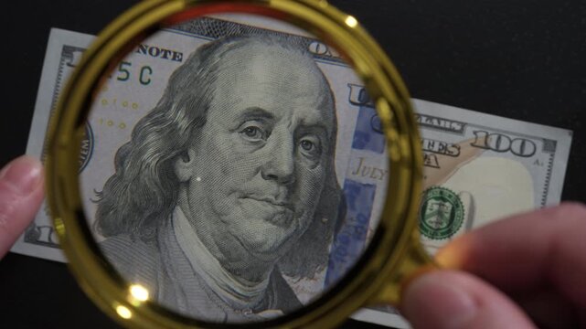Banker's hands examine banknote 100 dollar bill through magnifying glass, checking authenticity of cash and identifying counterfeits, visual control