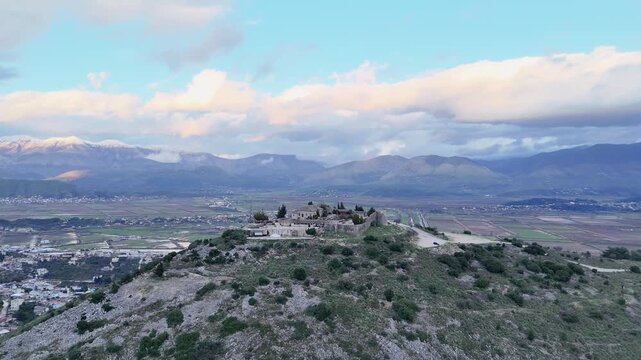 Aerial tracking shot moving forward over a rugged green hillside toward the hilltop Lekuresi Castle fortress, revealing a vast agricultural valley and snowy mountains in Albania